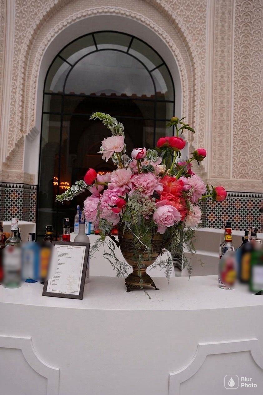Lush pink and coral peony arrangement in a brass urn on a bar in front of a Moroccan arched window and zellige tiling, floral design by SBAI Events Production