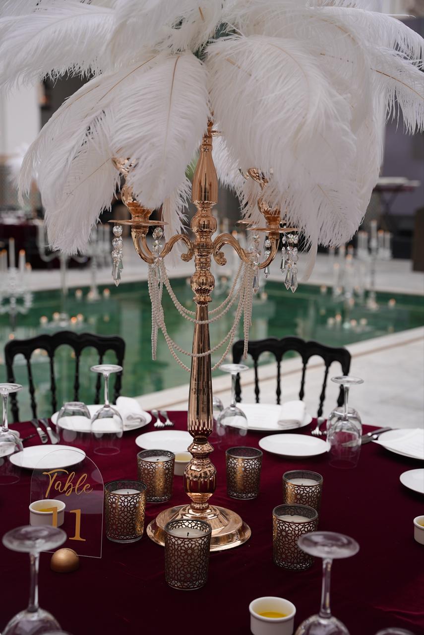 Rose-gold candelabra crowned with white ostrich feathers and pearl garlands on a burgundy velvet table, centerpiece by SBAI Events Production