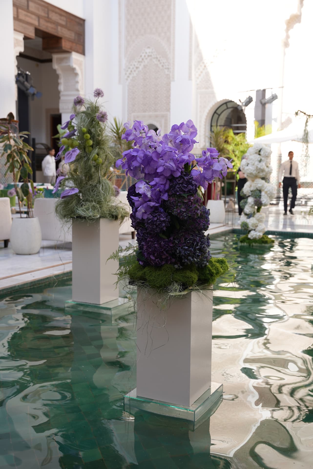 Gala Dinner Vegetal Garden at the patio of the Oberoi Marrakech — purple vanda orchids and anthuriums on white plinths above the emerald reflecting pool, ivory lounges and brass lanterns, produced by SBAI Events Production