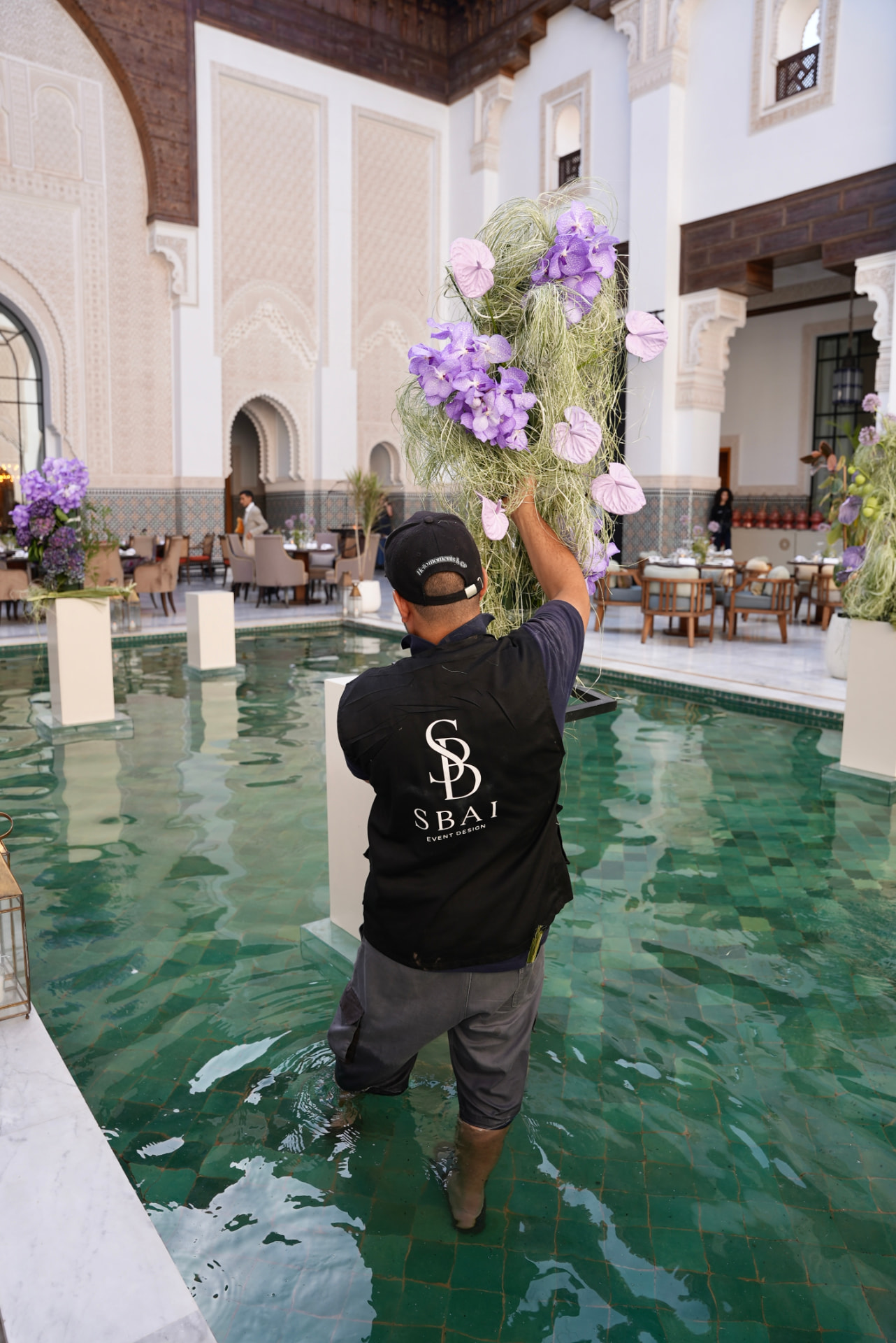 Gala Dinner Vegetal Garden at the patio of the Oberoi Marrakech — purple vanda orchids and anthuriums on white plinths above the emerald reflecting pool, ivory lounges and brass lanterns, produced by SBAI Events Production